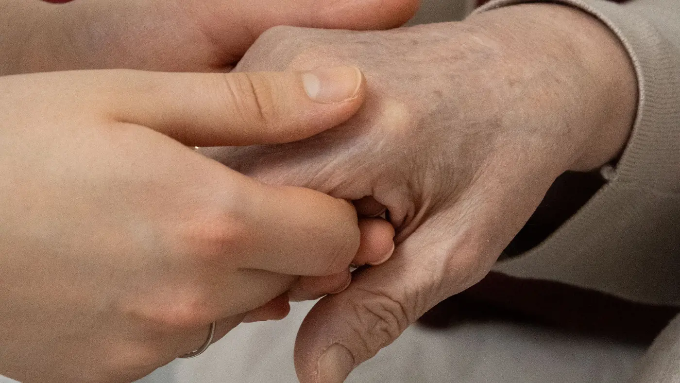 A younger person's hands gently holding an older person's hand, resting on a chair