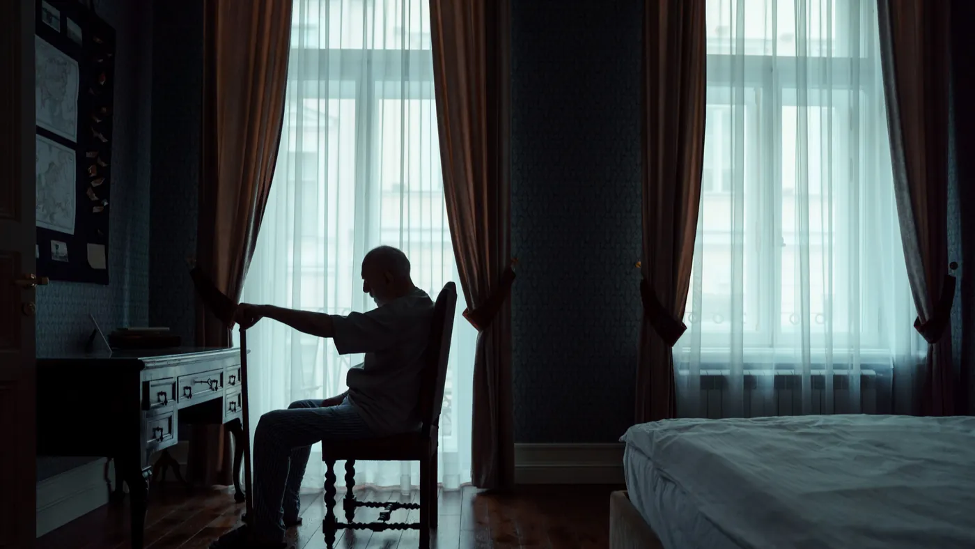 An older man sitting alone with a cane in a dim bedroom, looking toward a curtained window beside an empty bed